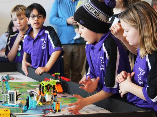 Photo by Sophia Lord. A group of elementary aged youth in matching purple shirts surround a robot on a table.